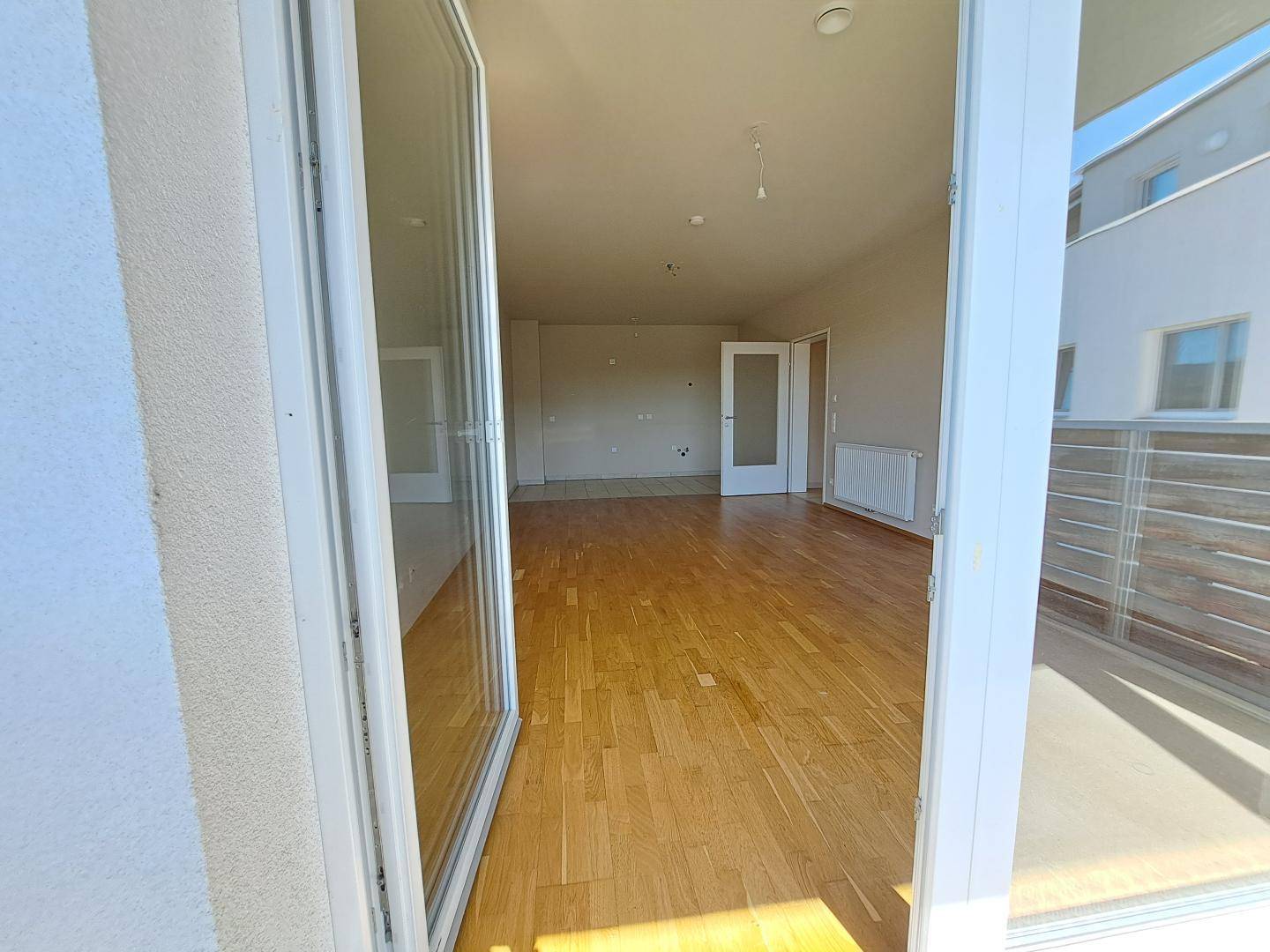 Blick vom Balkon in einen hellen, leeren Wohnbereich mit Holzboden und viel Tageslicht.