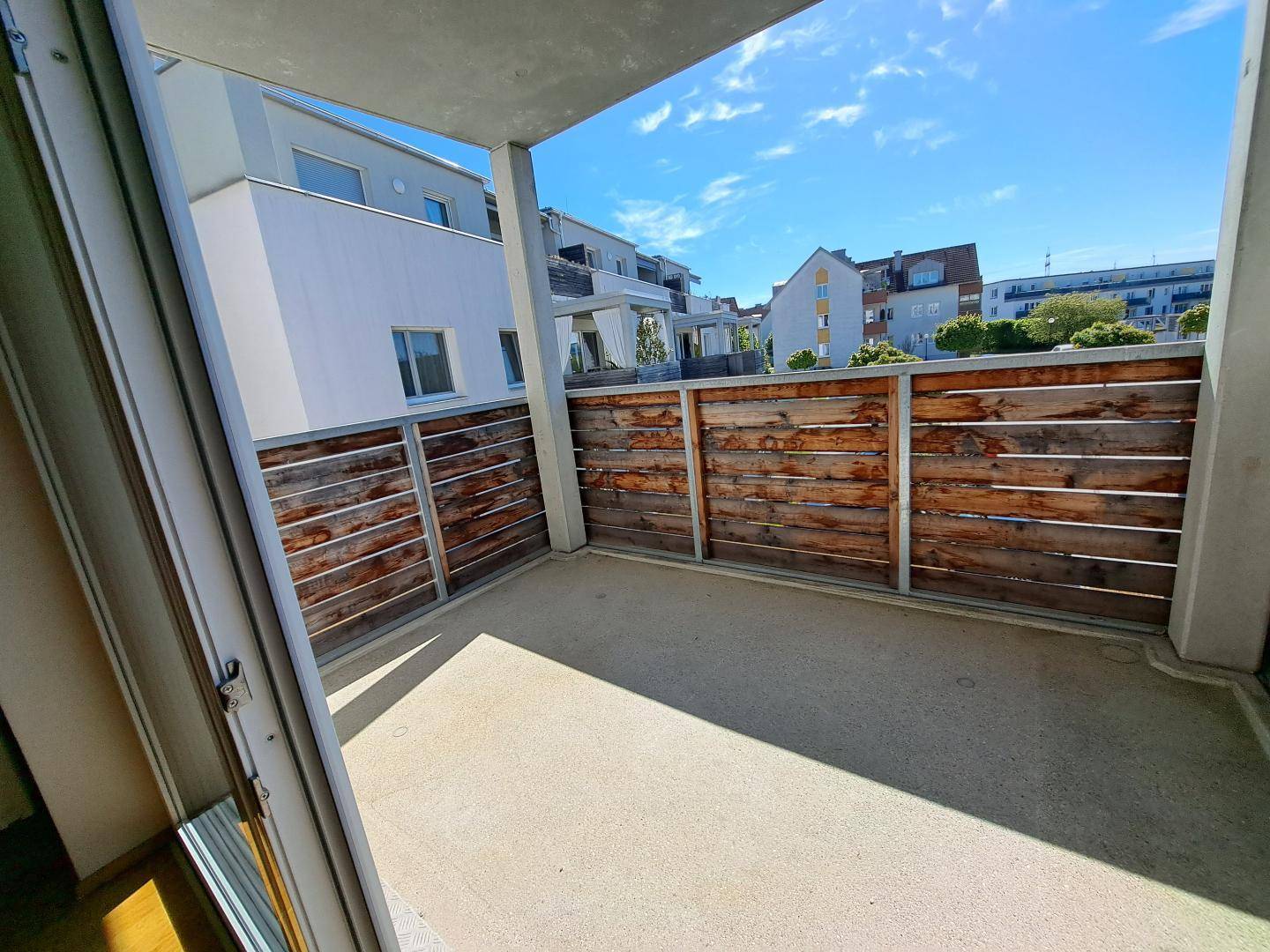 Sonniger Balkon mit Holzsichtschutz und Blick auf die moderne Wohnanlage unter blauem Himmel.