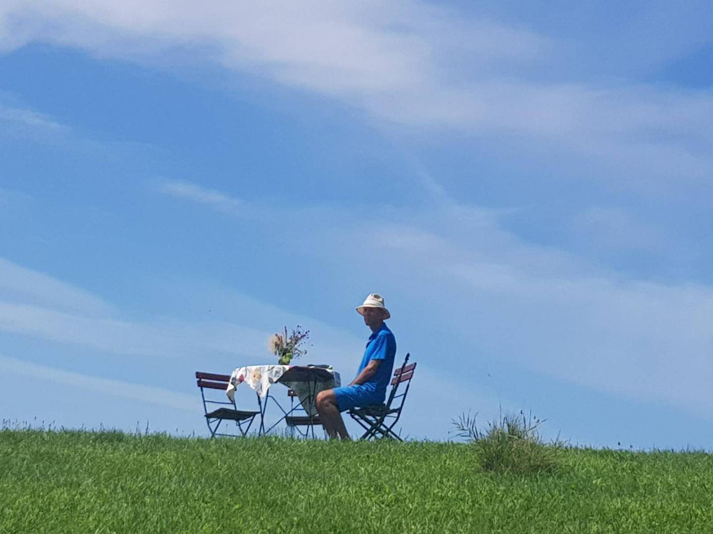 Eine Person sitzt an einem Tisch auf einer weiten grünen Wiese unter einem klaren blauen Himmel.