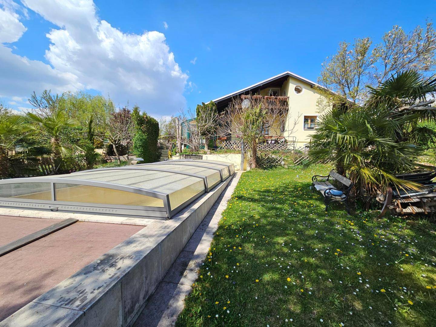 Großer Garten mit überdachtem Pool und dem Haus im Hintergrund unter blauem Himmel.