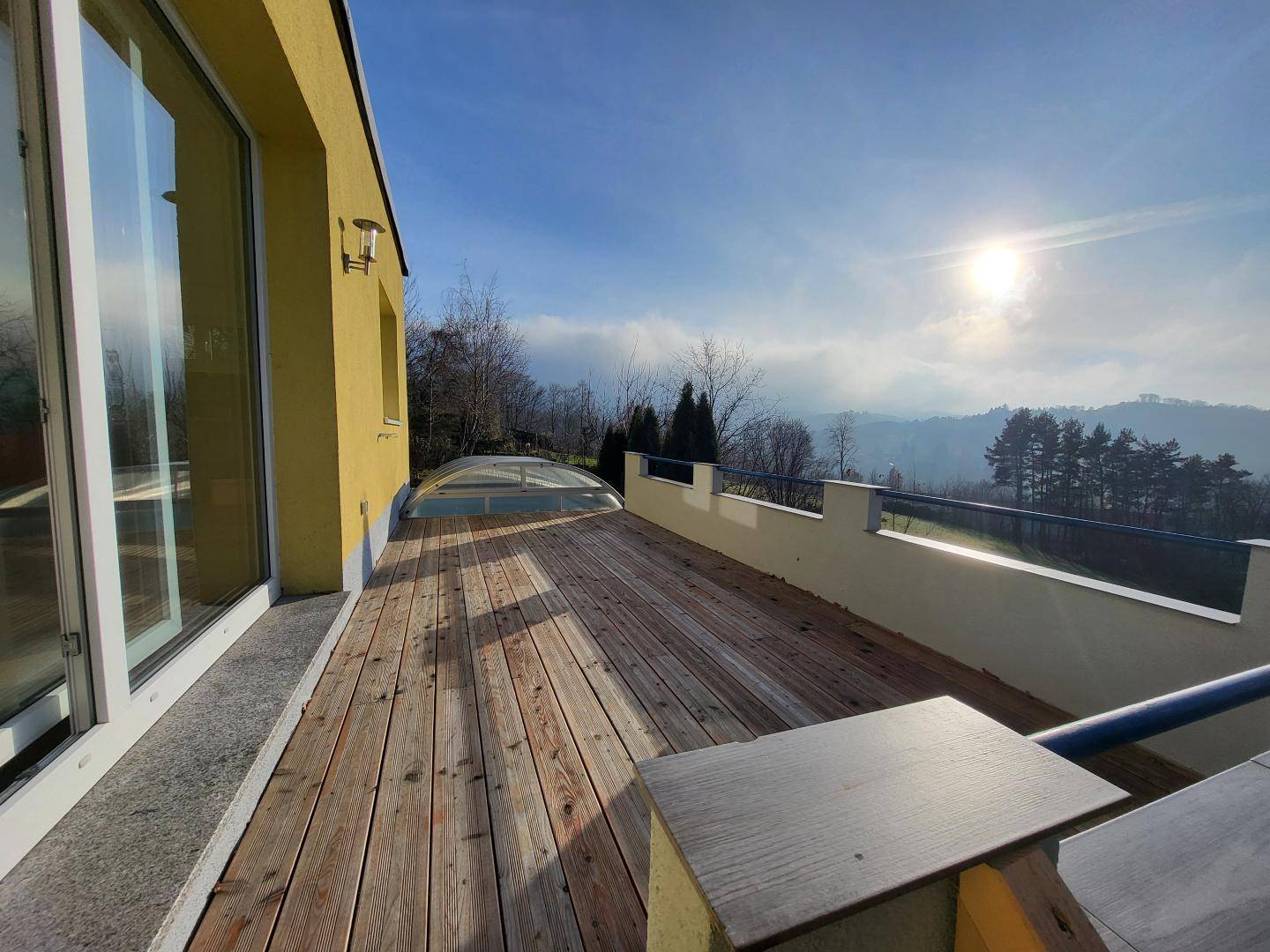 Sonnige Holzterrasse mit weitem Blick über die Landschaft und den überdachten Poolbereich.