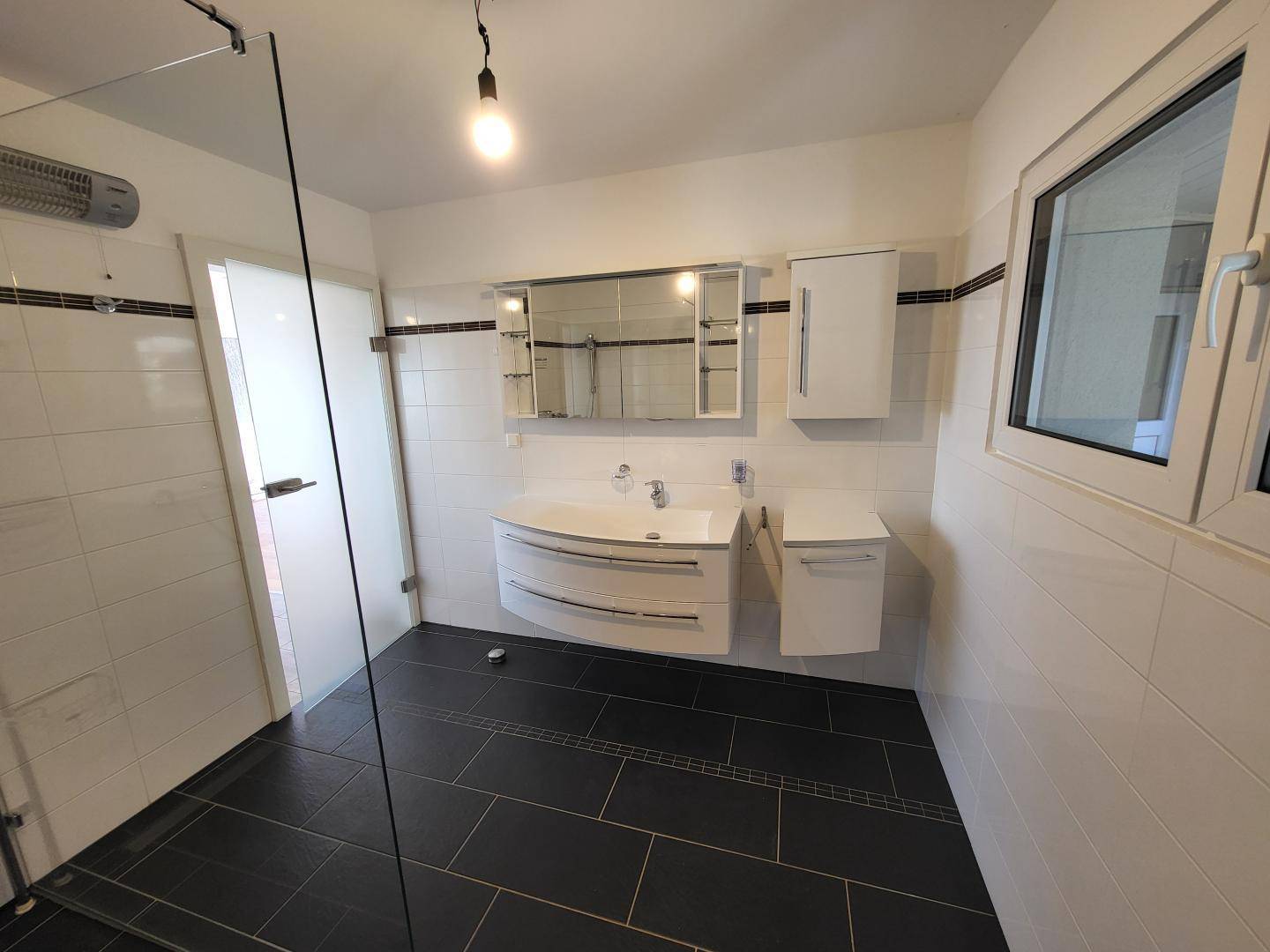 Modernes Badezimmer mit dunklen Fliesen, großer Dusche und Fenster für Tageslicht.