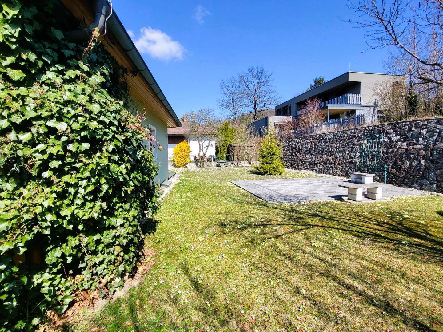 Seitlicher Blick auf den weitläufigen Garten mit Rasenfläche, einer gepflasterten Terrasse und einer Steinmauer im Hintergrund.