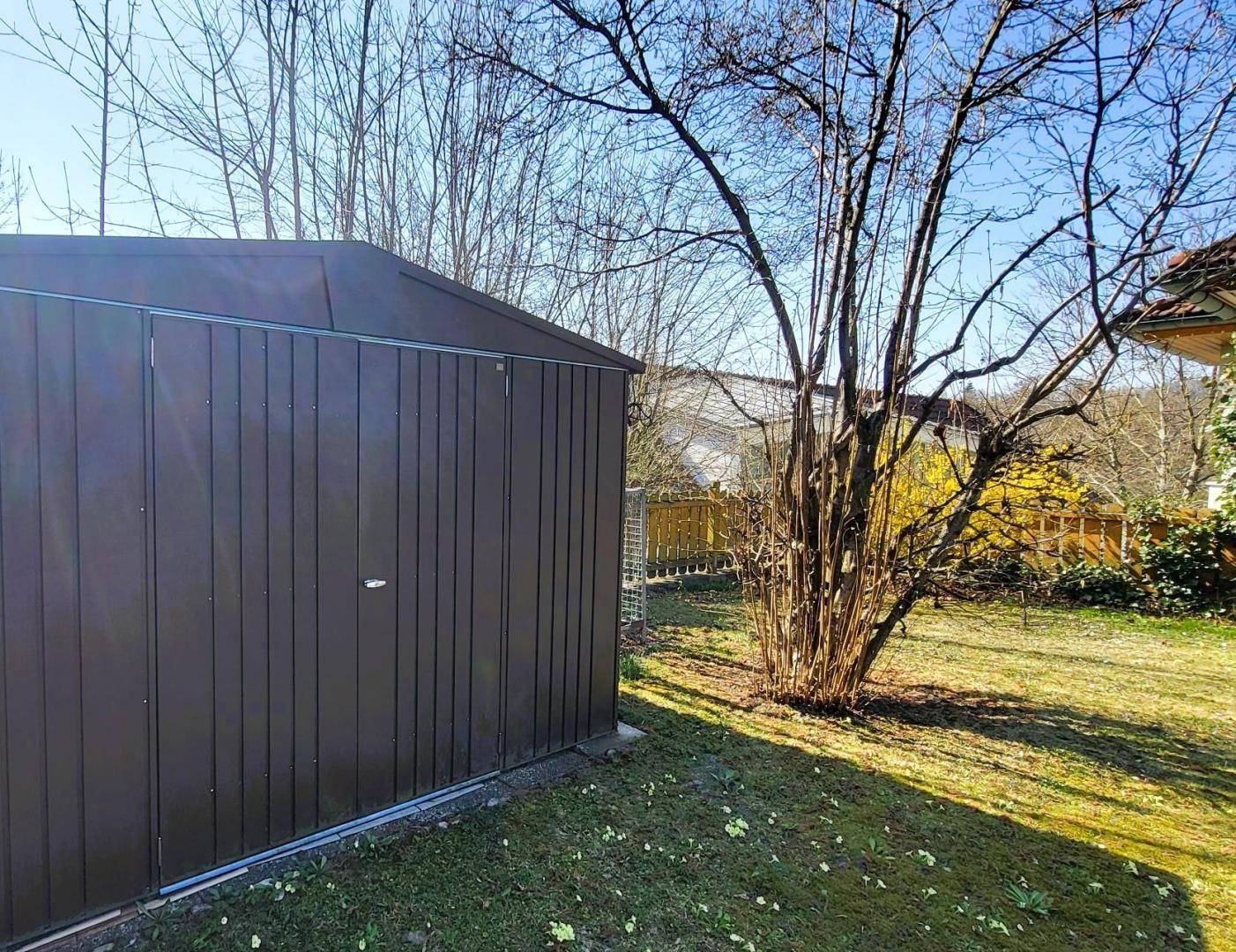 Praktischer Gartenschuppen aus Metall, umgeben von Bäumen und Sträuchern in einem sonnigen Garten.