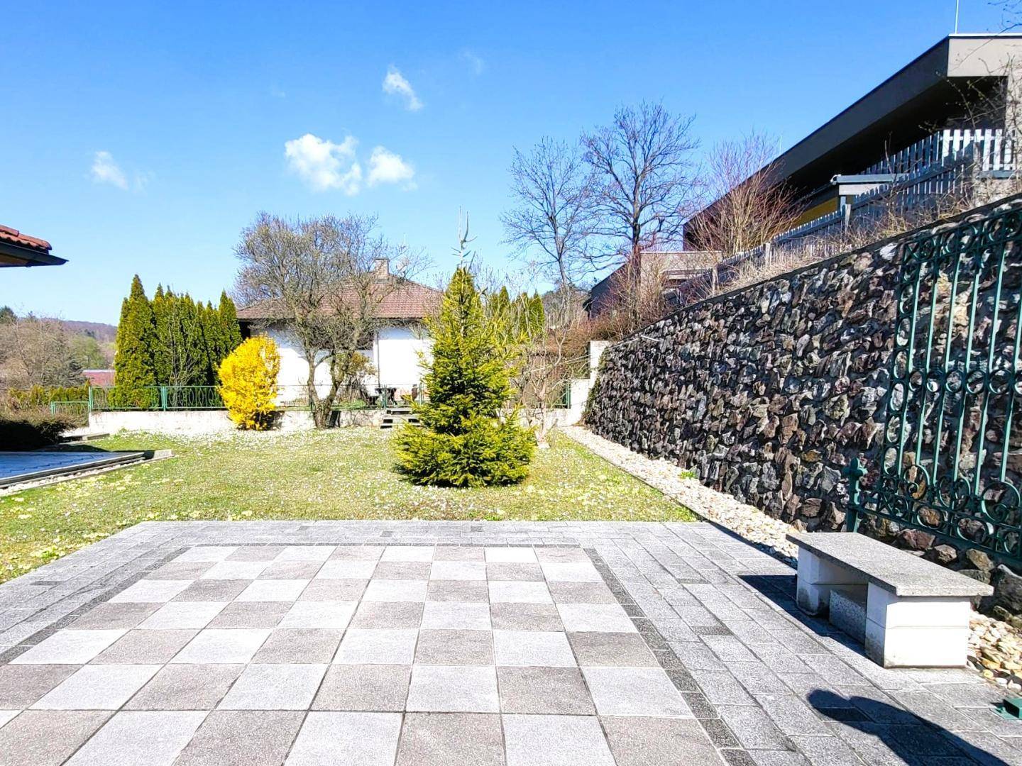 Große gepflasterte Terrasse im Garten mit einer Steinbank, umgeben von Grünflächen und einer Steinmauer.