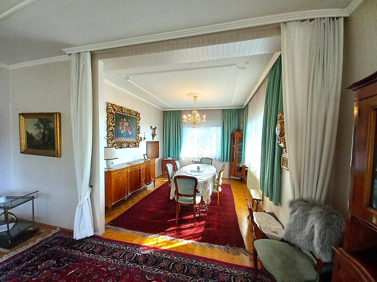 Eleganter Essbereich mit großem Esstisch, antiken Holzmöbeln, grünen Vorhängen und Kronleuchter.