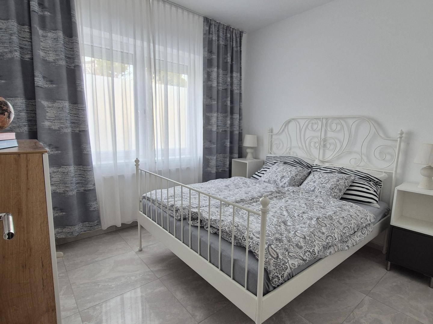 Helles Schlafzimmer mit weißem Metallbett, gemusterten Vorhängen und Fliesenboden.