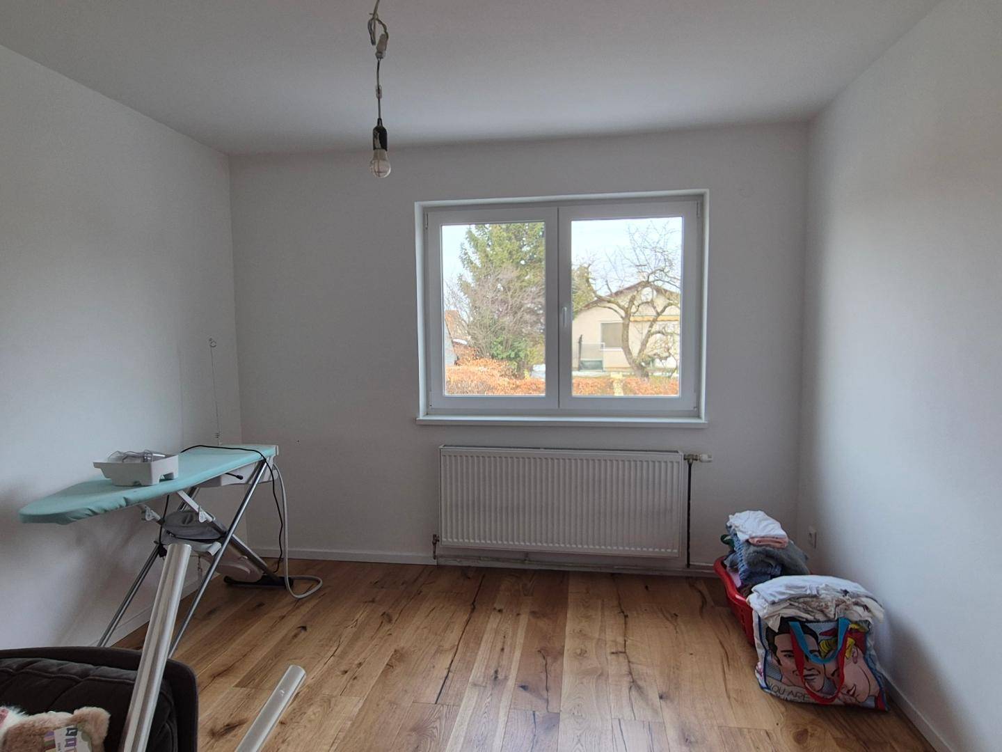 Leeres Zimmer mit Holzboden, weiß gestrichenen Wänden und großem Fenster.