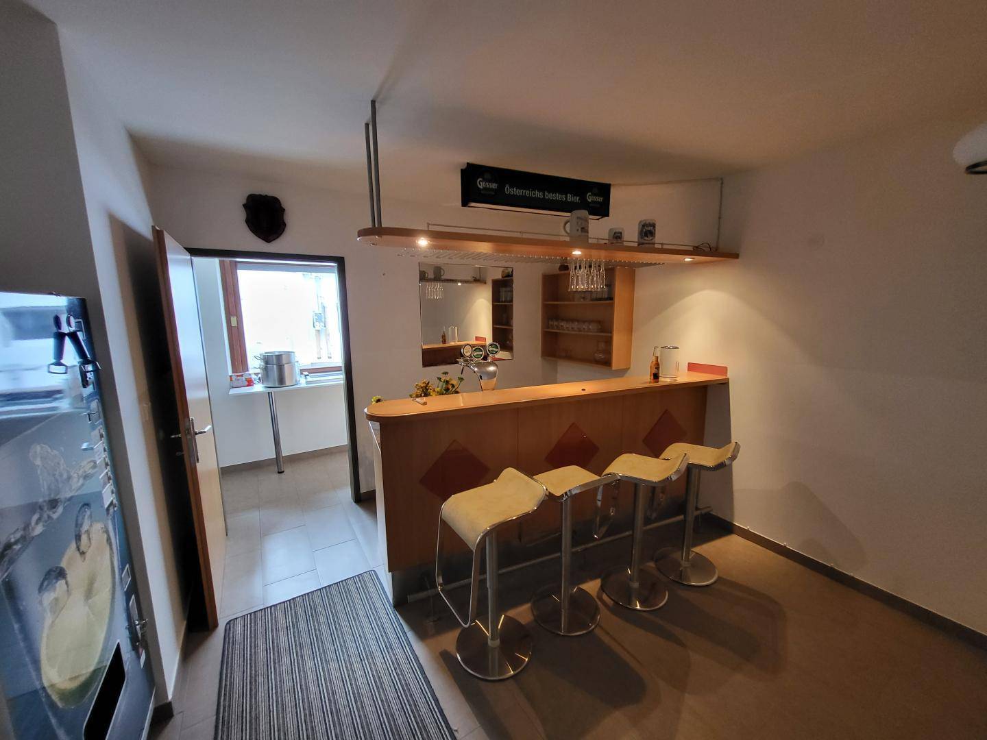 Kleine Bar mit Holztresen, Barhockern und einem Fenster, das Tageslicht hereinlässt.