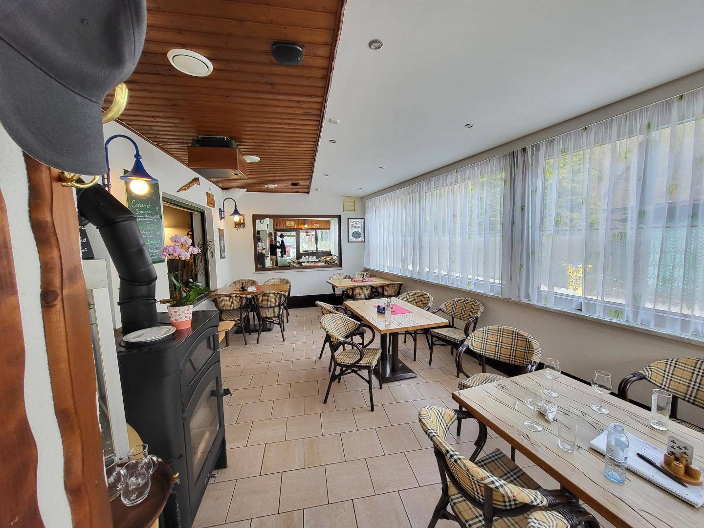 Heller Restaurantbereich mit Kaminofen, Holzbalkendecke und großen Fenstern mit hellen Vorhängen.