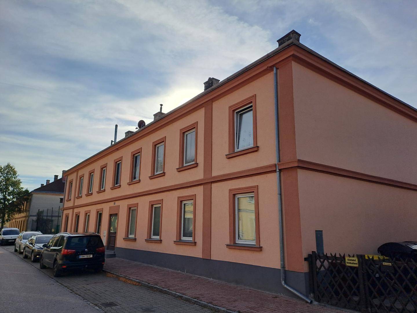 Straßenansicht des Gebäudes mit rosafarbener Fassade, vielen Fenstern und parkenden Autos davor.