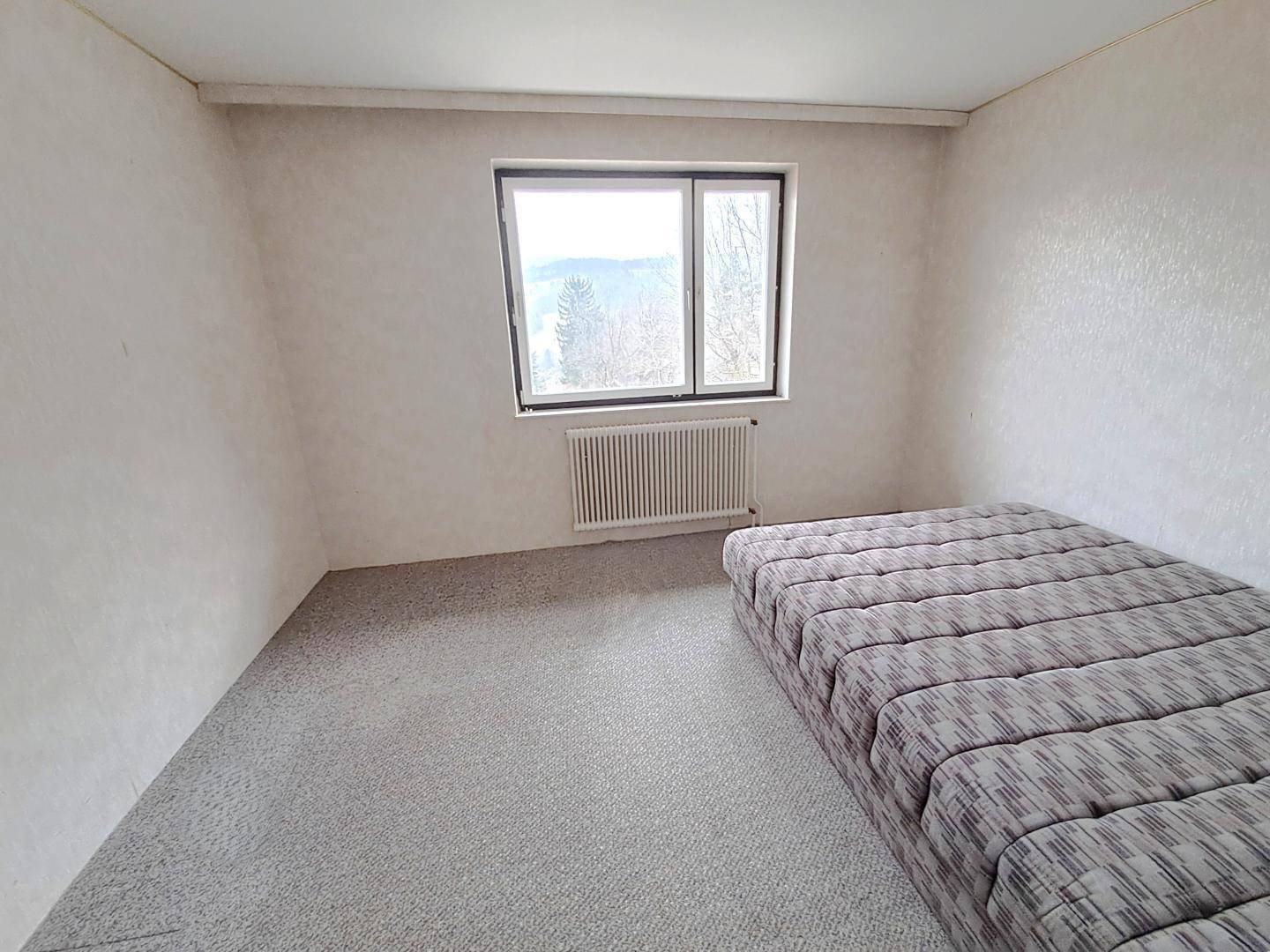 Schlafzimmer mit Teppichboden, großem Fenster und einfacher Einrichtung.