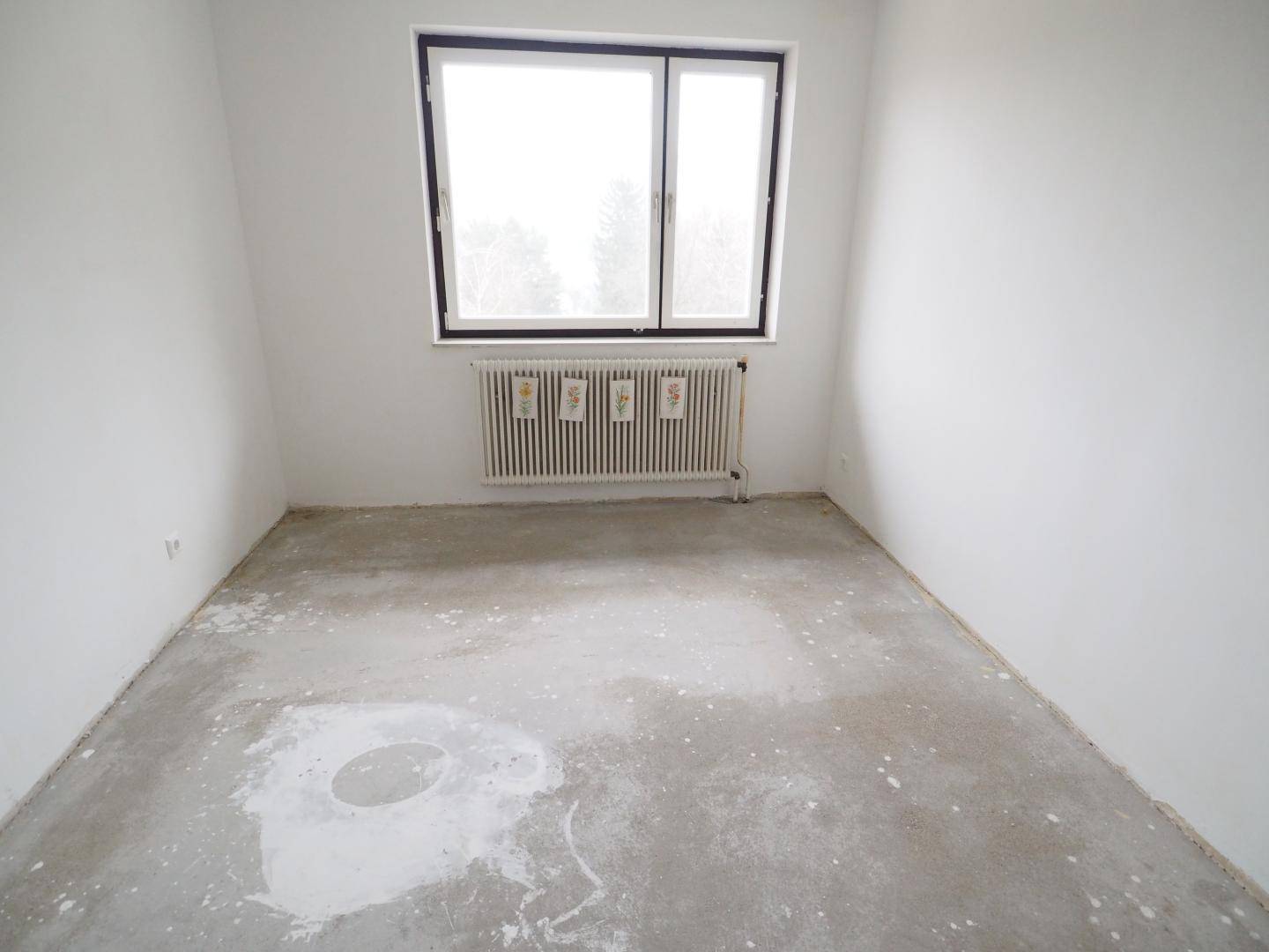 Leeres Zimmer mit Betonboden und großem Fenster, ideal für eine individuelle Gestaltung.
