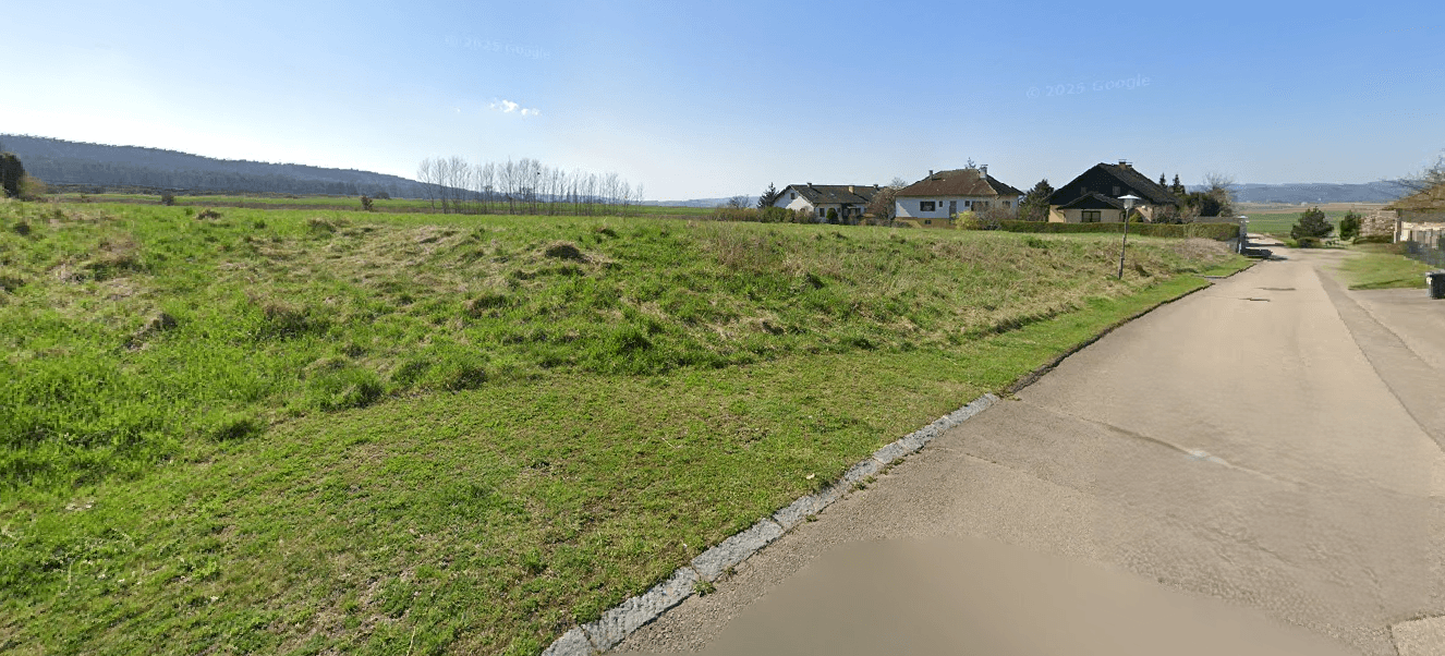 Grundstück neben einer Straße mit angrenzenden Häusern und weitem Blick in die grüne Umgebung.