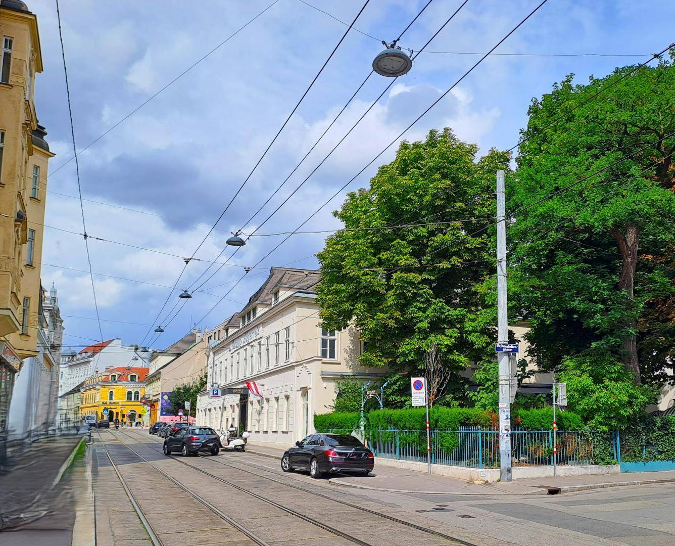 Städtische Straße mit Straßenbahnschienen, parkenden Autos und Gebäuden unter bewölktem Himmel.