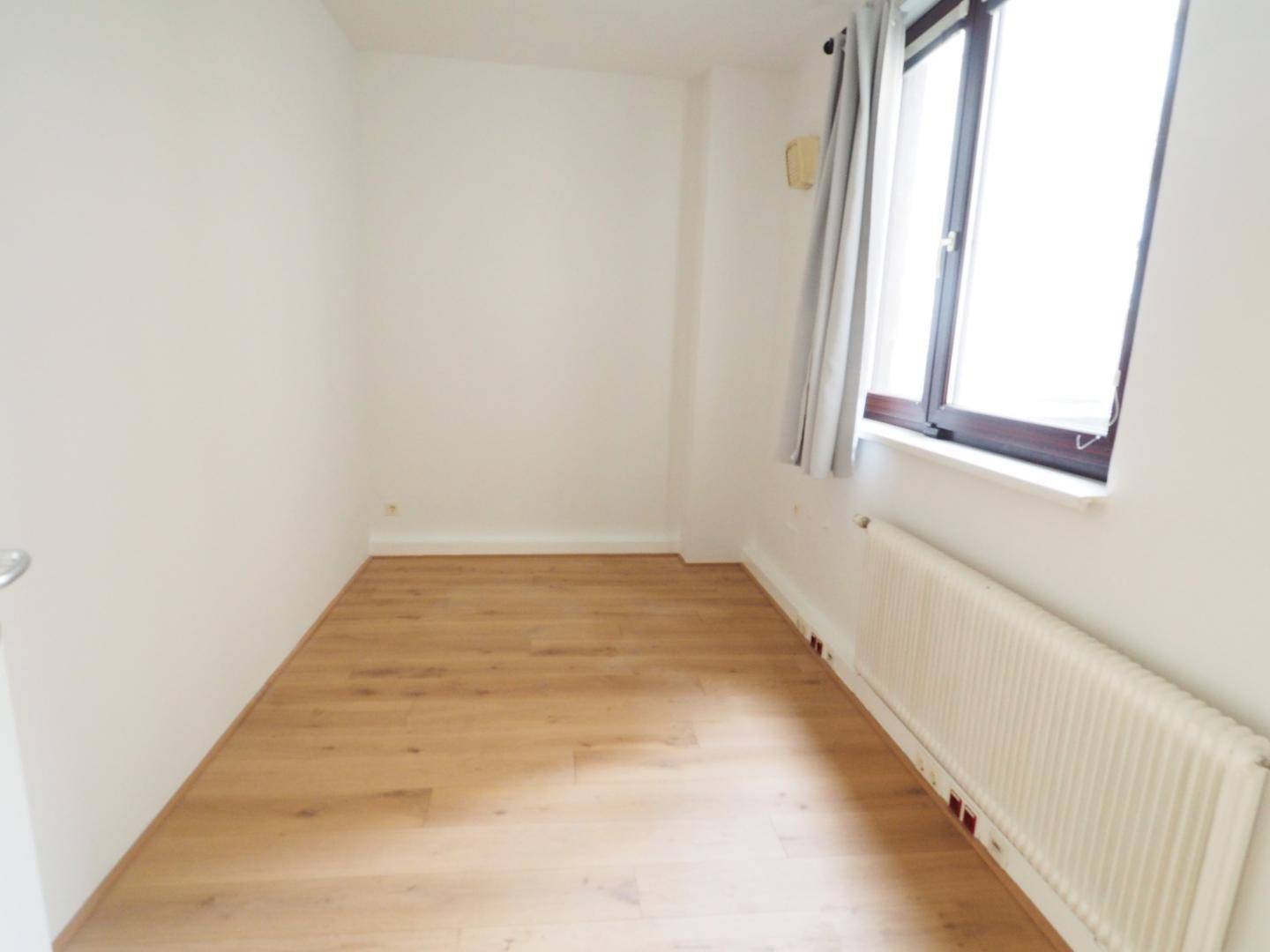 Helles Zimmer mit Holzboden, weißen Wänden und einem Fenster mit Vorhängen und Heizkörper.