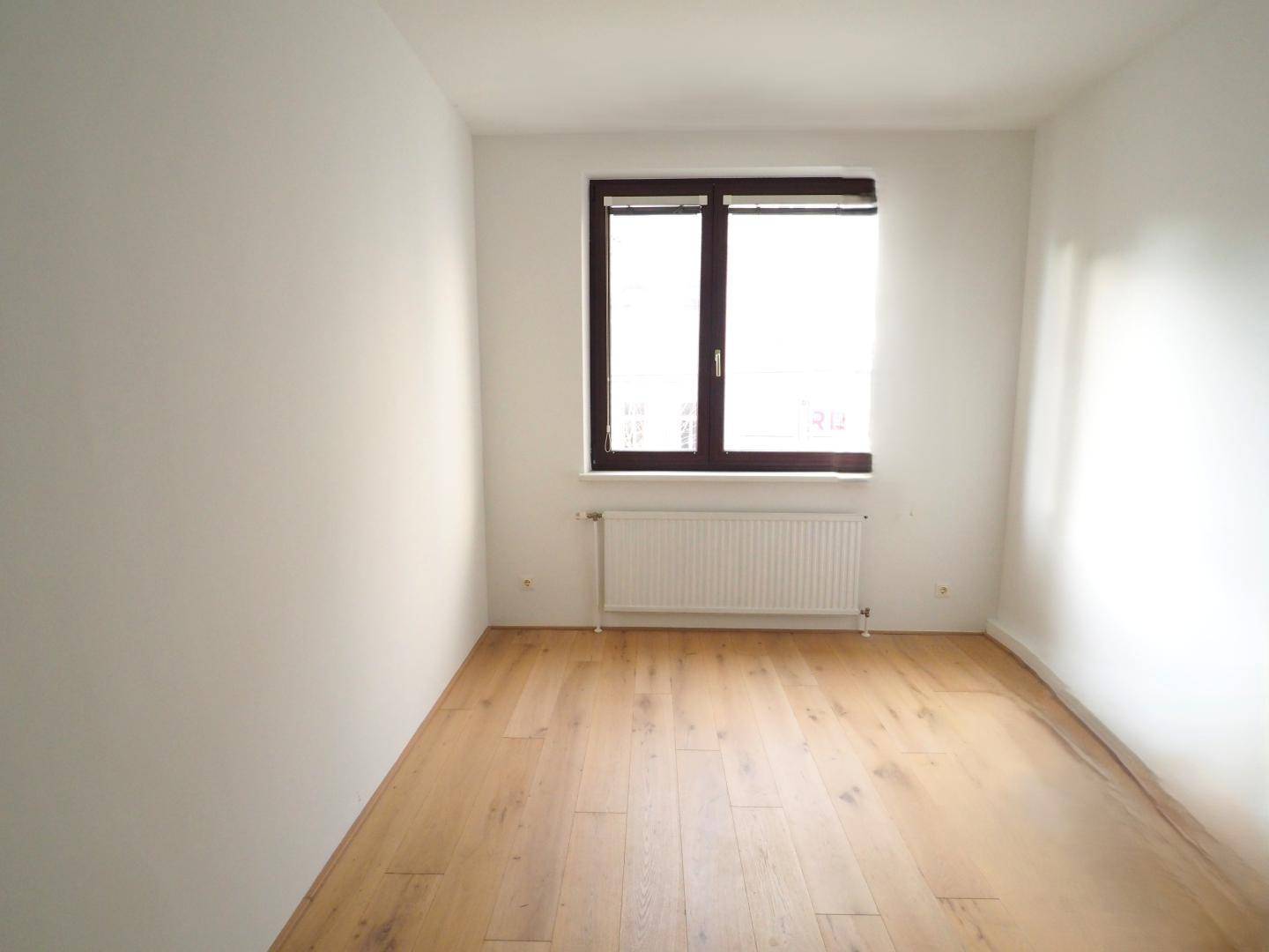 Schlafzimmer mit hellem Holzboden, weißen Wänden und einem Fenster mit Heizkörper darunter.