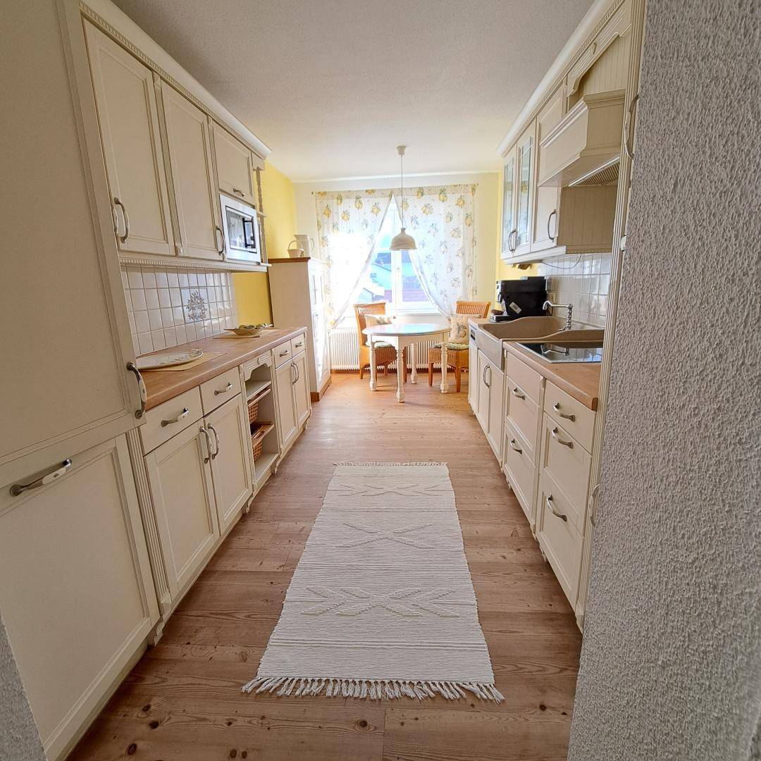Helle Küche mit weißen Schränken, Holzboden und einem Essbereich am Fenster.