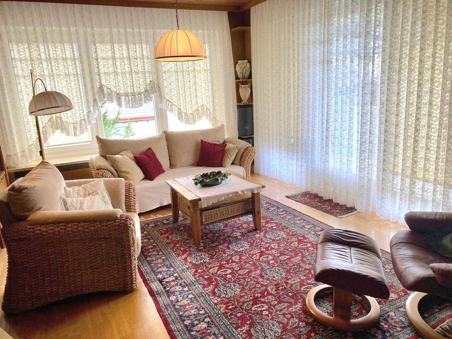 Gemütliches Wohnzimmer mit Korbmöbeln, Teppich und großen Fenstern mit Vorhängen.