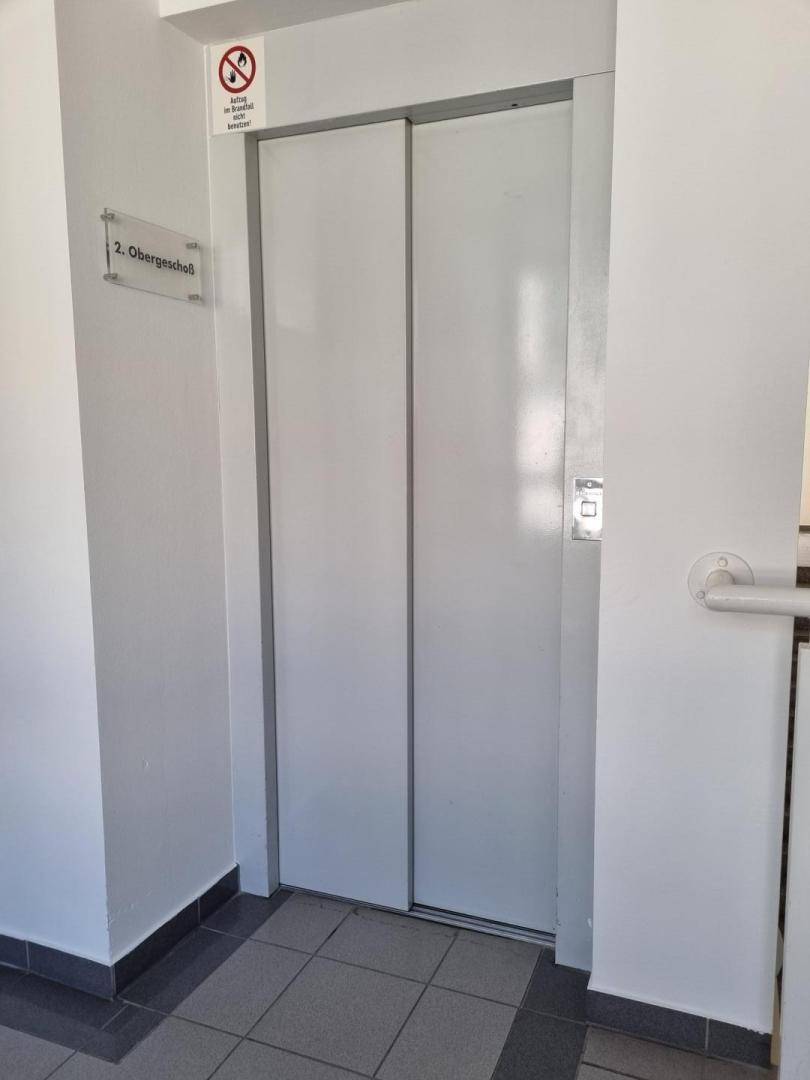 Aufzugstür in einem hellen Flur mit Fliesenboden, der zum zweiten Obergeschoss führt.