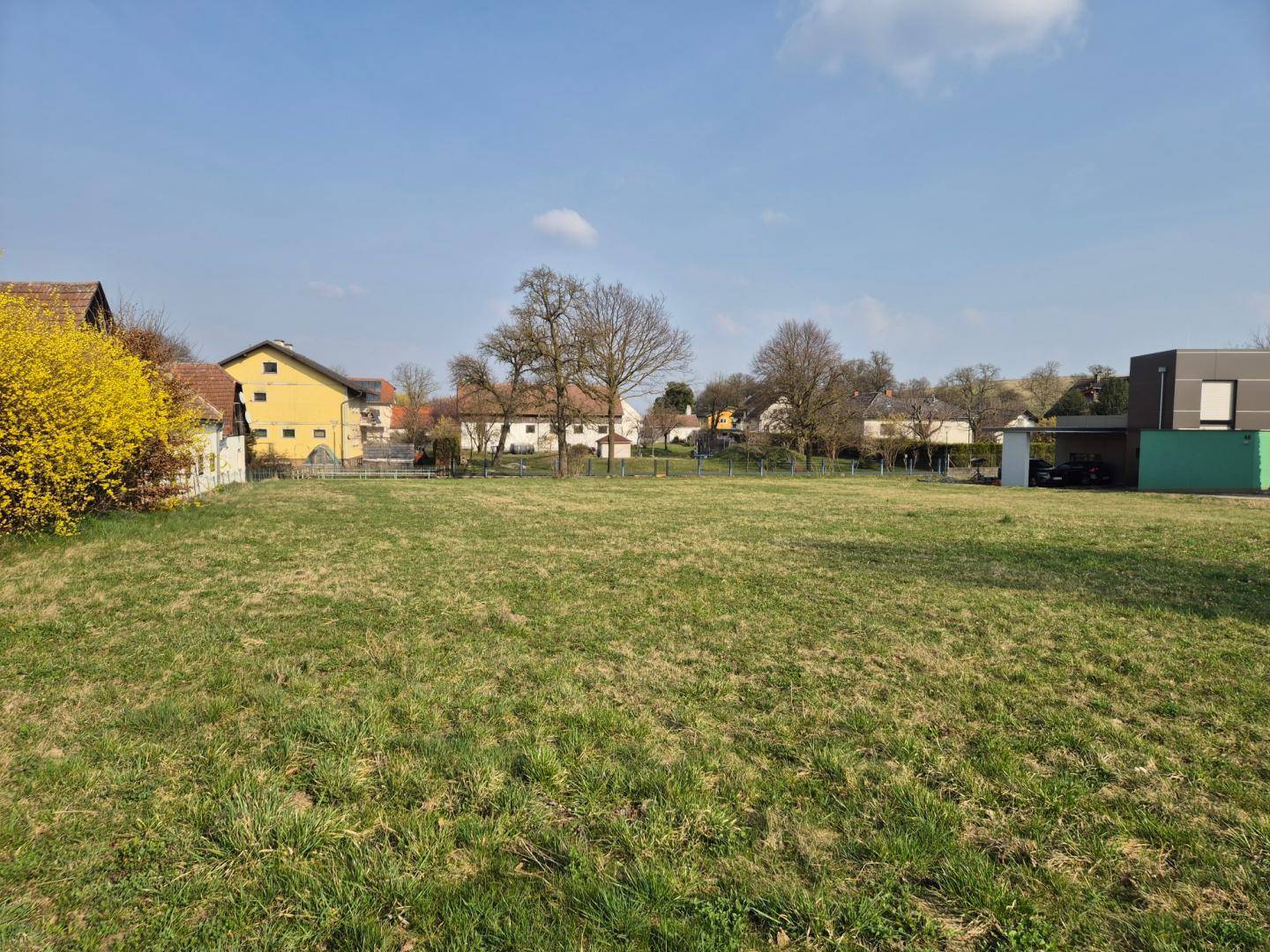 Großes, offenes Wiesengrundstück unter blauem Himmel, mit Wohngebäuden und Bäumen in der Ferne.
