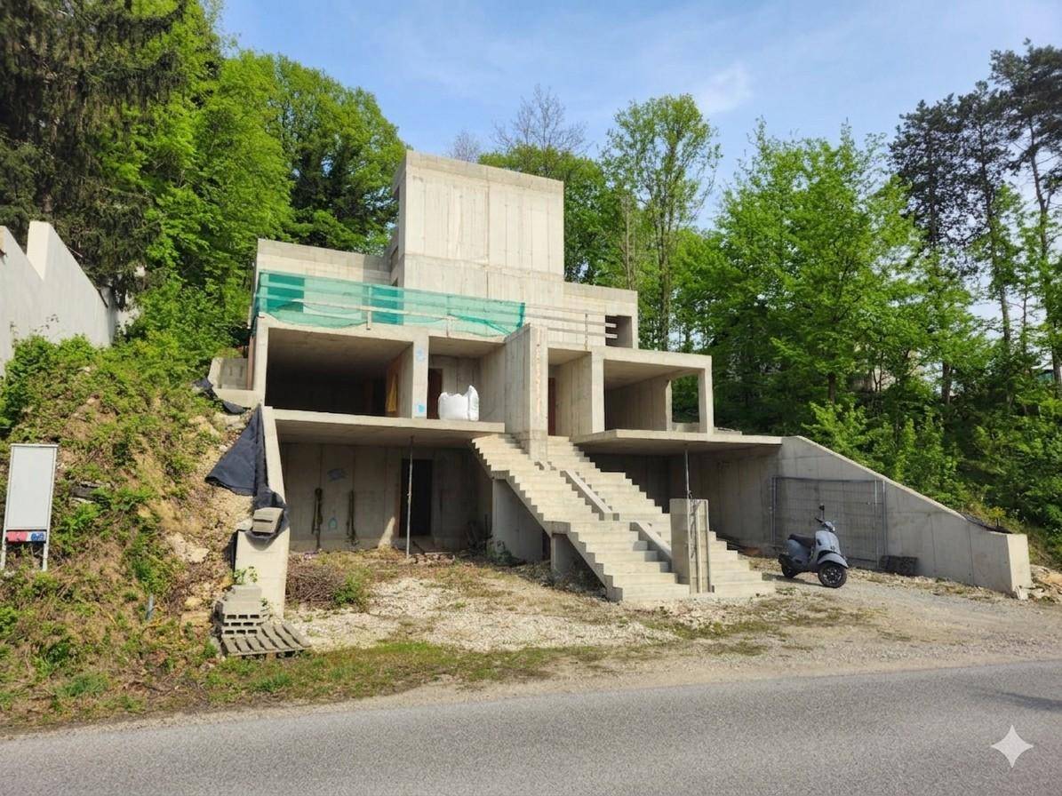 Rohbau einer modernen Villa mit Betonstruktur und Treppen, umgeben von Bäumen und einer Straße.