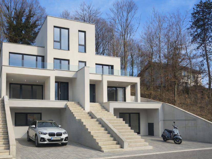 Elegante moderne Villa mit weißer Fassade, großen Fenstern und Glasgeländern, eingebettet in eine hügelige Landschaft.