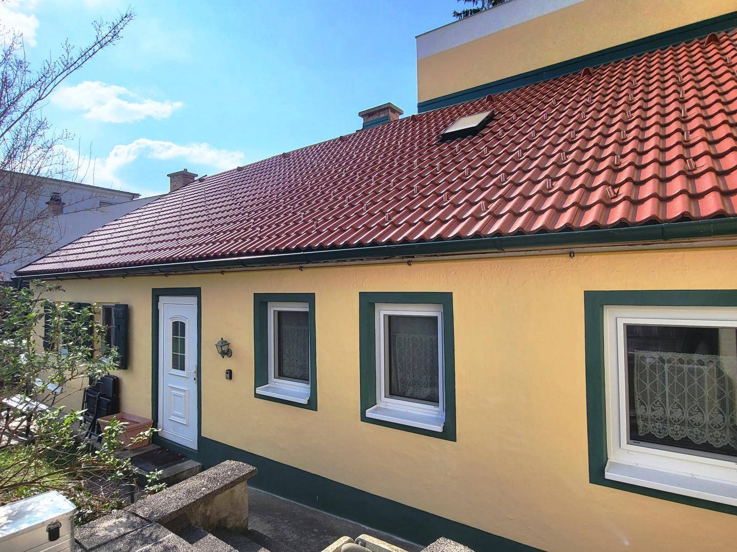 Seitenansicht des Hauses mit gelber Fassade, weißer Eingangstür und rotem Ziegeldach.