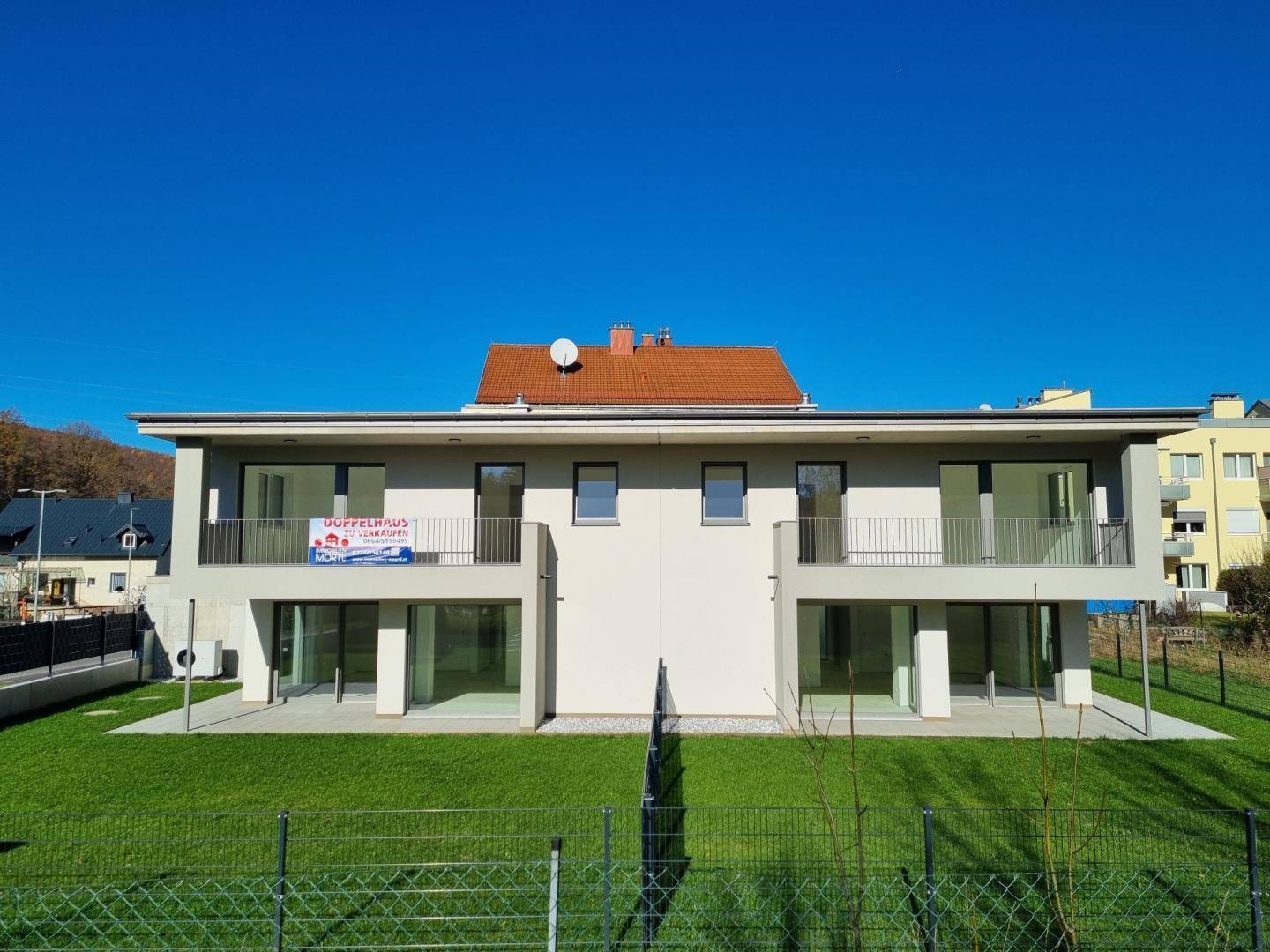 Vorderansicht eines modernen Doppelhauses mit großem Garten und Balkonen unter blauem Himmel.
