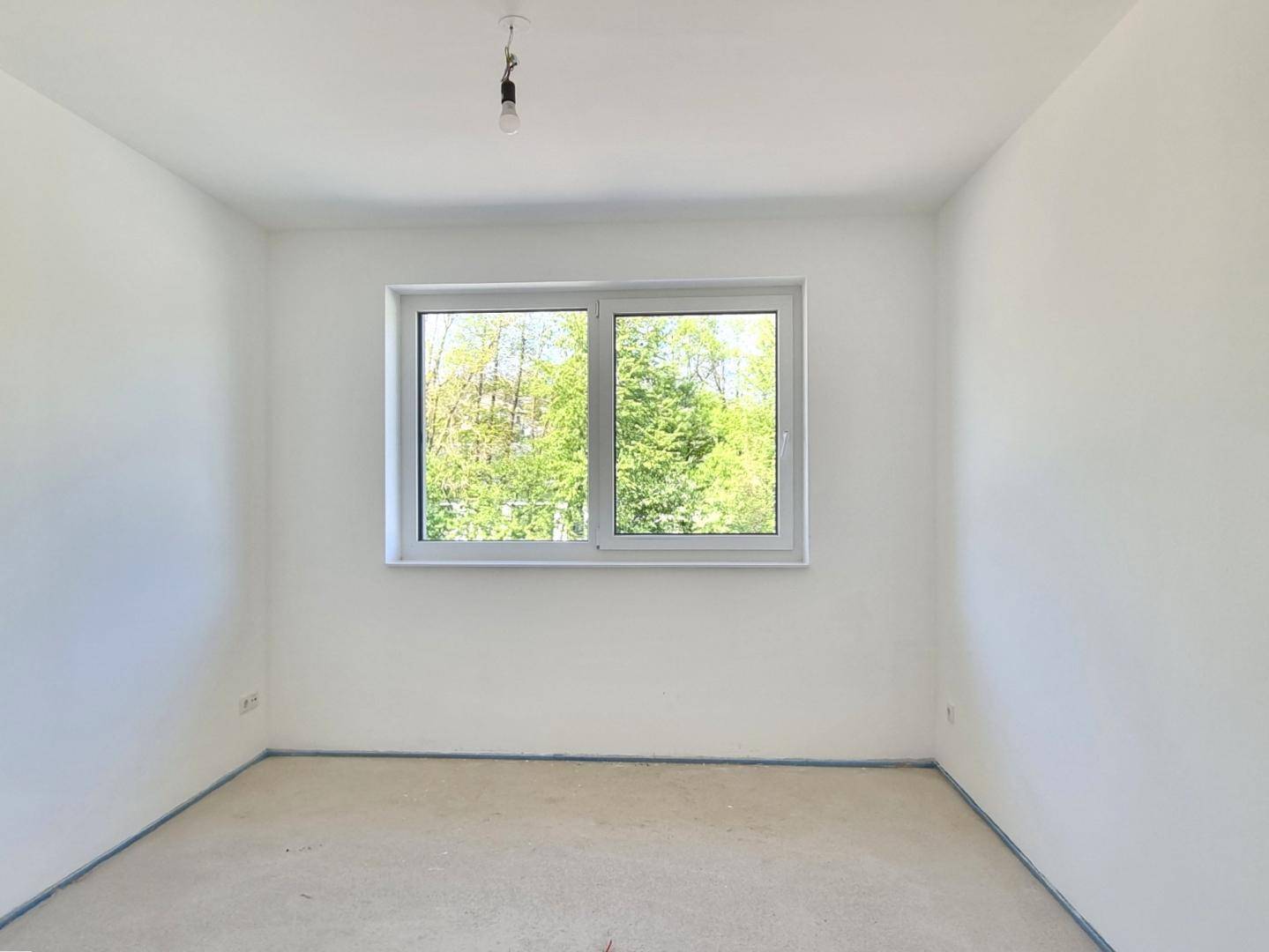 Heller Raum im Rohbau mit großem Fenster und Blick auf die grüne Umgebung.
