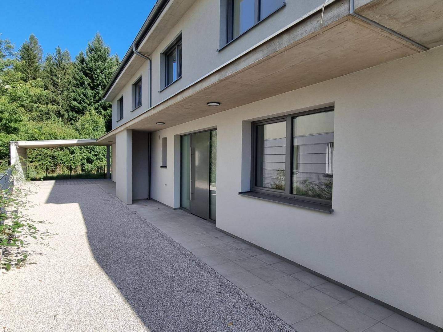 Seitenansicht des modernen Gebäudes mit Carport und Zufahrt, umgeben von Bäumen.
