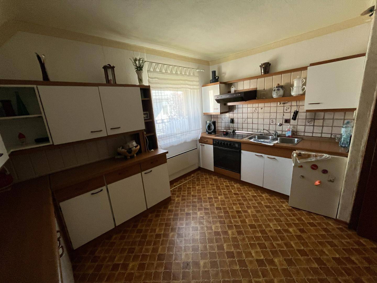 Ältere Küche mit gemustertem Fliesenboden, weißen und braunen Schränken und Fenster.