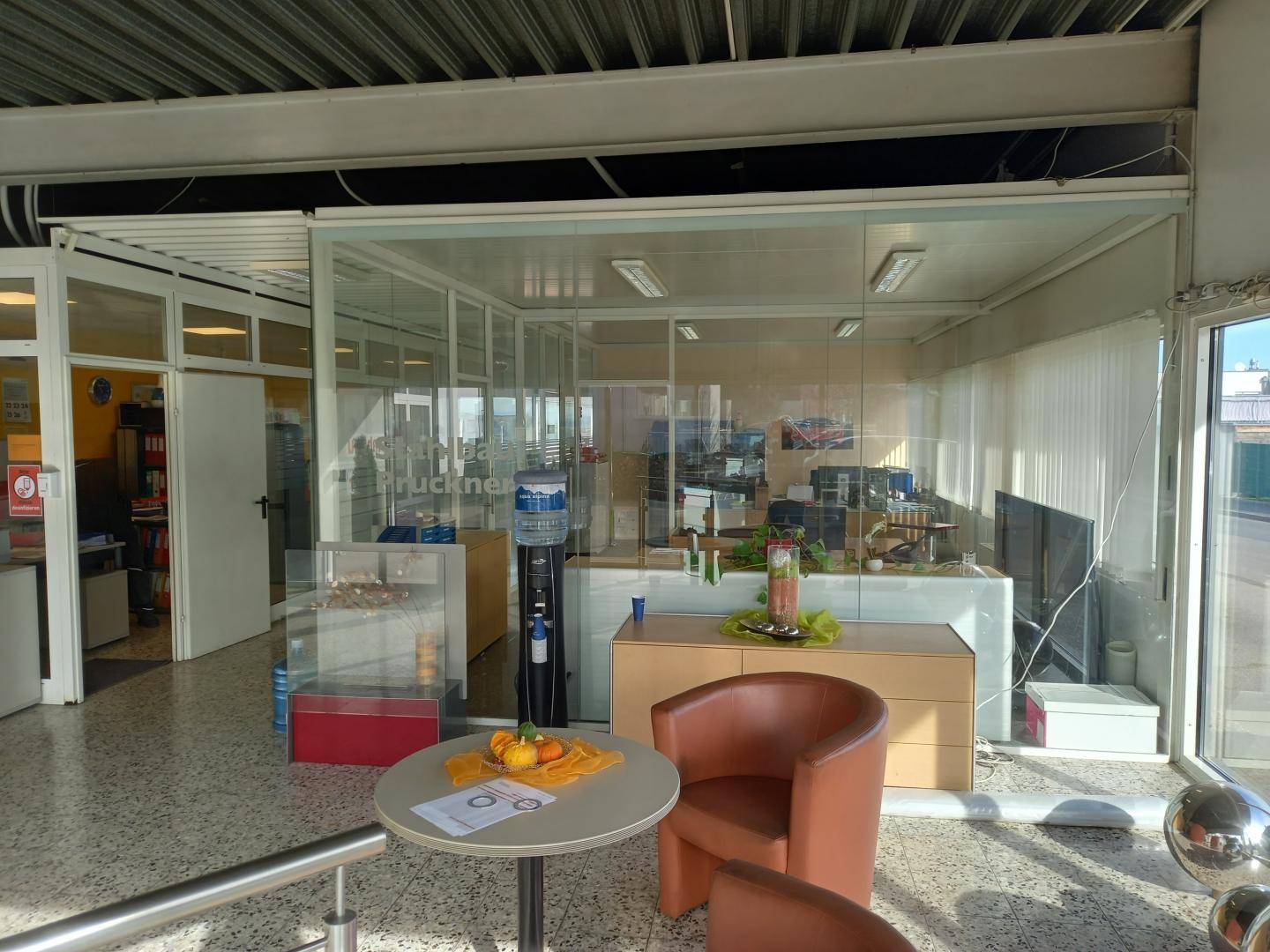 Empfangsbereich mit Sitzgelegenheiten, einem kleinen Tisch und einem durch Glaswände abgetrennten Bürobereich im Hintergrund.