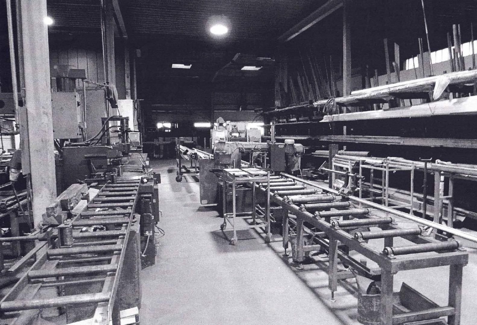 Schwarz-Weiß-Aufnahme einer Werkstatt oder Produktionshalle mit verschiedenen Maschinen und Lagerregalen für Materialien.