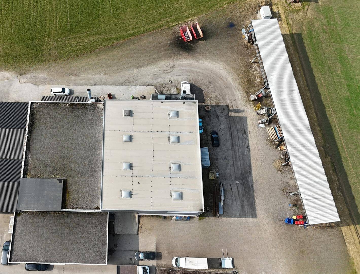 Luftaufnahme eines Industrieareals mit mehreren Gebäuden, einer langen Überdachung und angrenzenden Feldern.