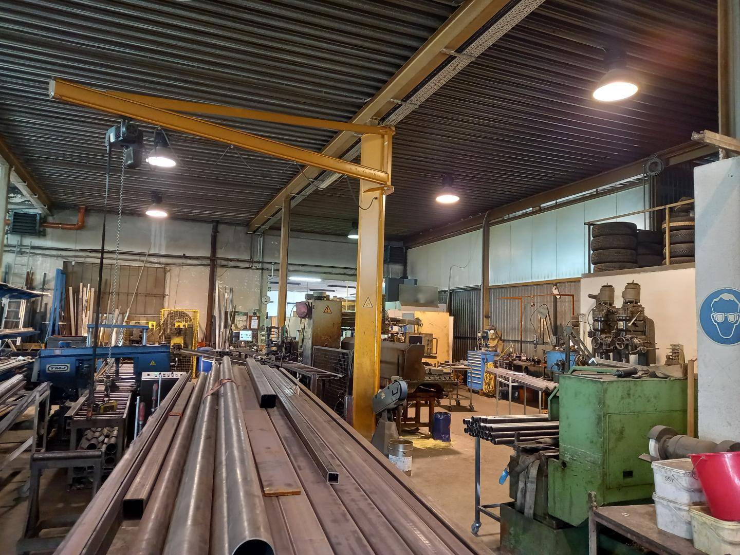 Farbaufnahme einer Werkstatt mit Metallrohren, verschiedenen Maschinen und einem gelben Schwenkkran.