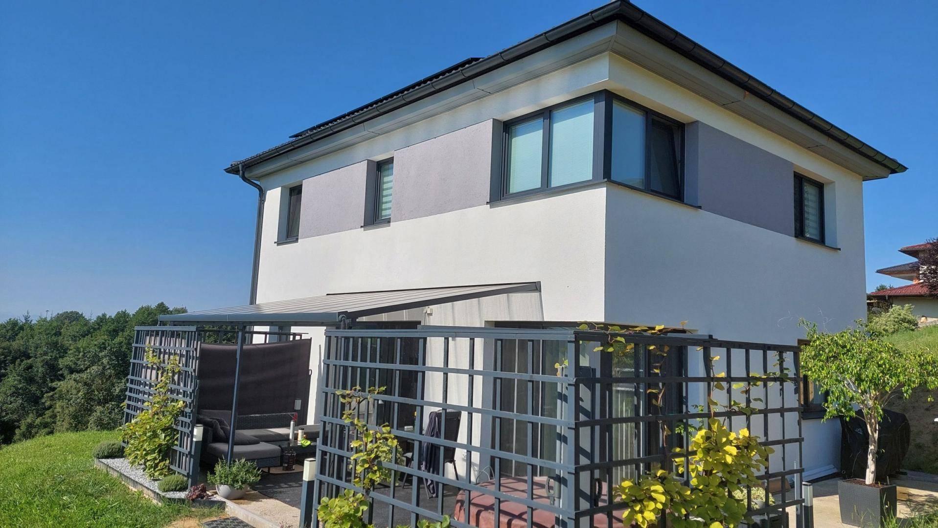 Modernes Wohnhaus mit überdachter Terrasse und Blick ins Grüne unter blauem Himmel.