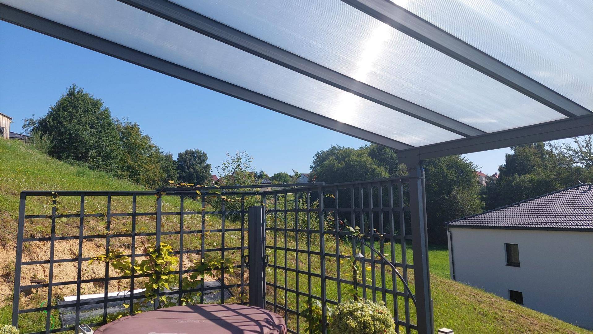Überdachte Terrasse mit transparentem Dach und Gitterwerk, umgeben von grünen Hügeln.