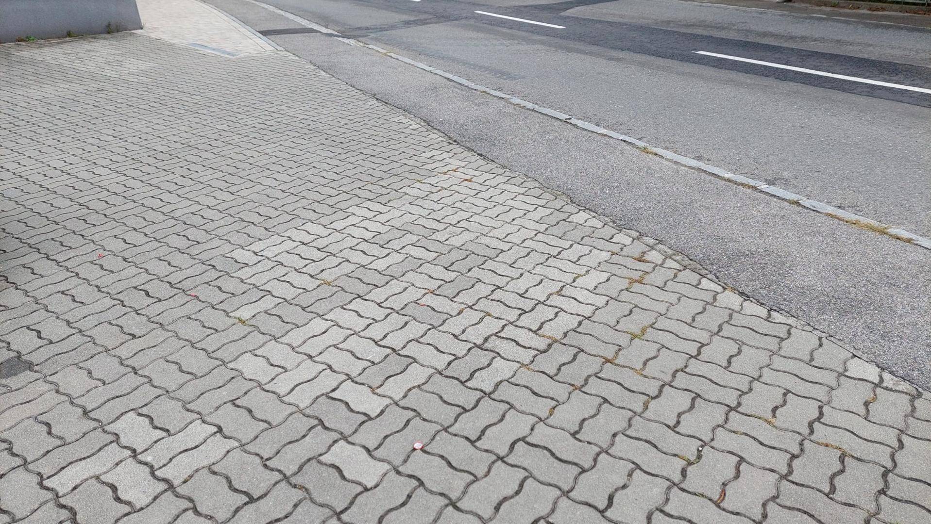 Gepflasterter Gehweg neben einer asphaltierten Straße mit Fahrbahnmarkierungen unter hellem Himmel.