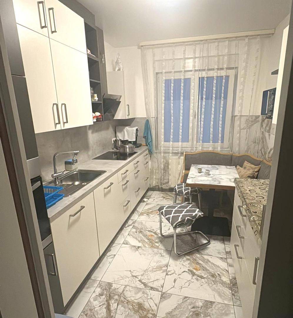 Moderne Küche mit weißen Schränken, Fliesenboden und kleinem Essbereich am Fenster.