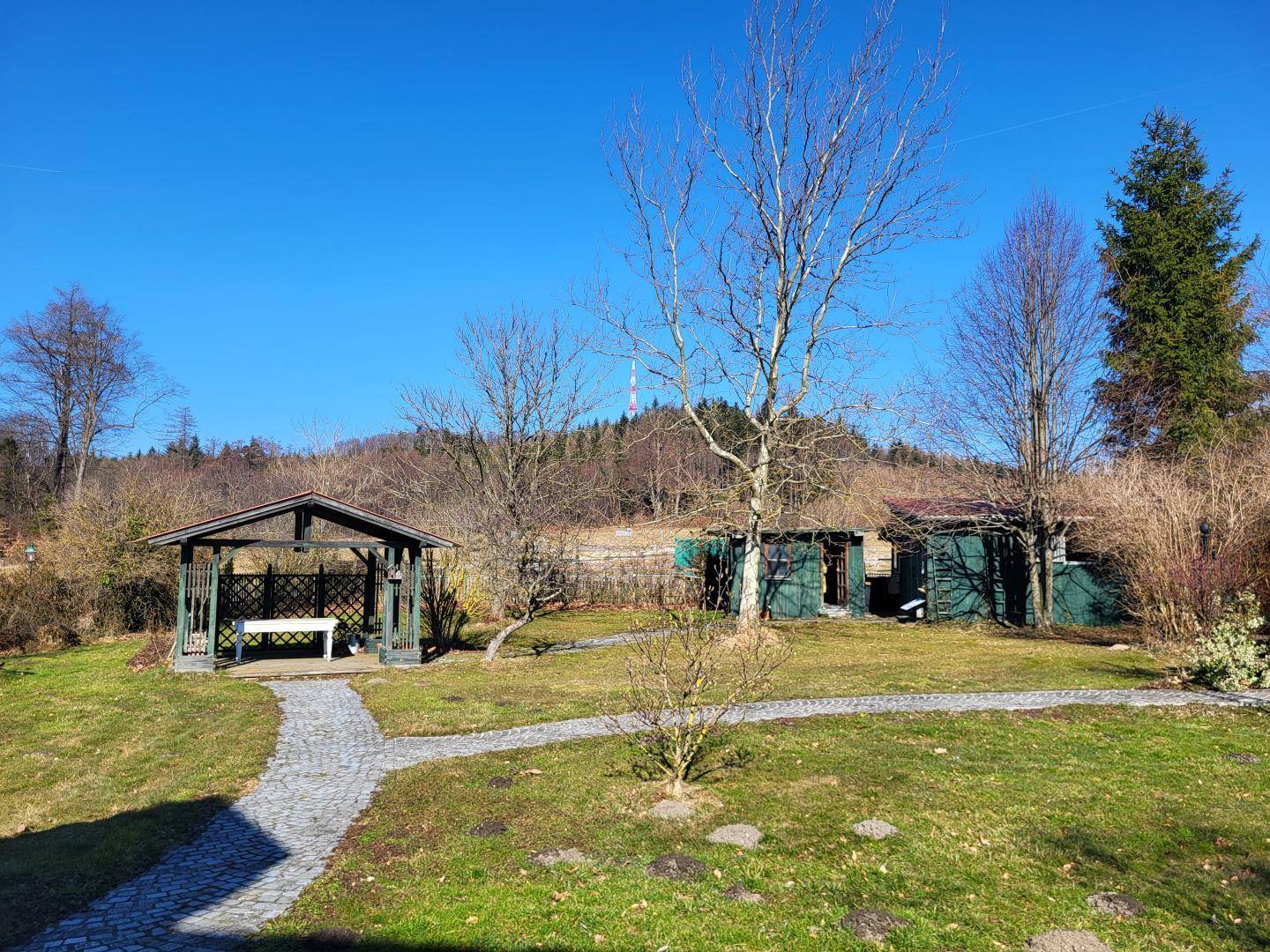 Gartenbereich mit einem Holzpavillon, gepflastertem Weg und kleinen Schuppen im Hintergrund, umgeben von Bäumen.