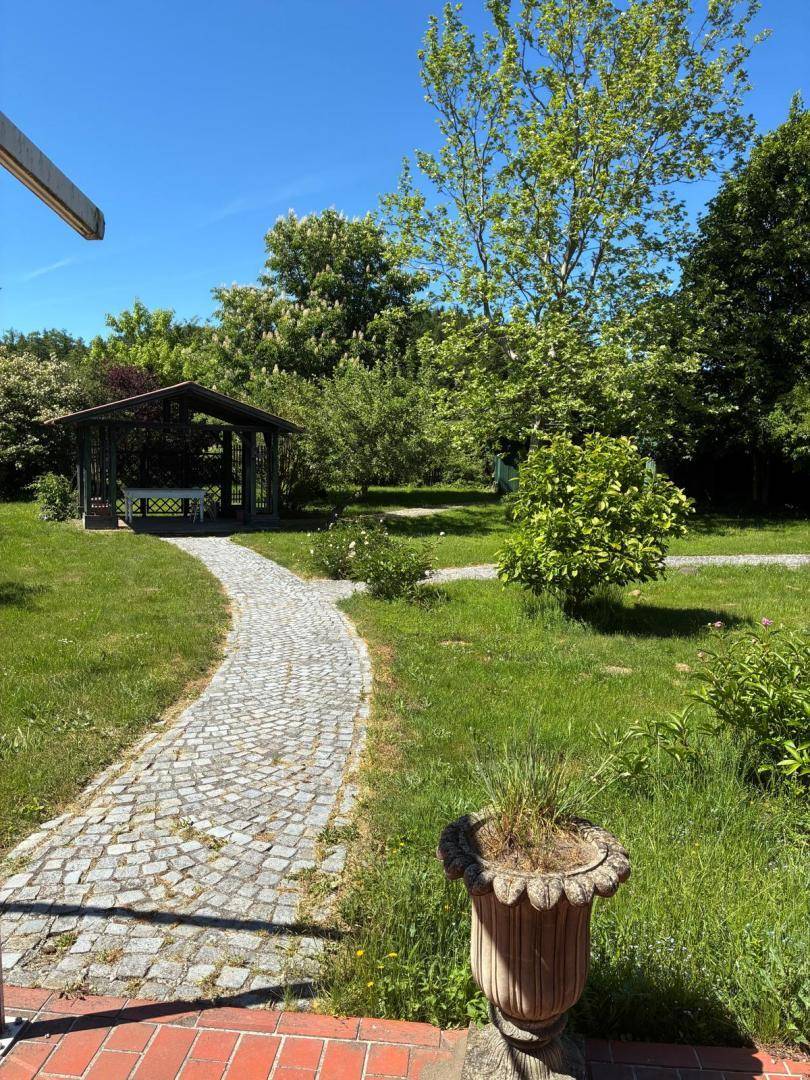 Gartenansicht von der Terrasse mit gepflastertem Weg, grünem Rasen, Büschen und einem Pavillon in der Ferne.
