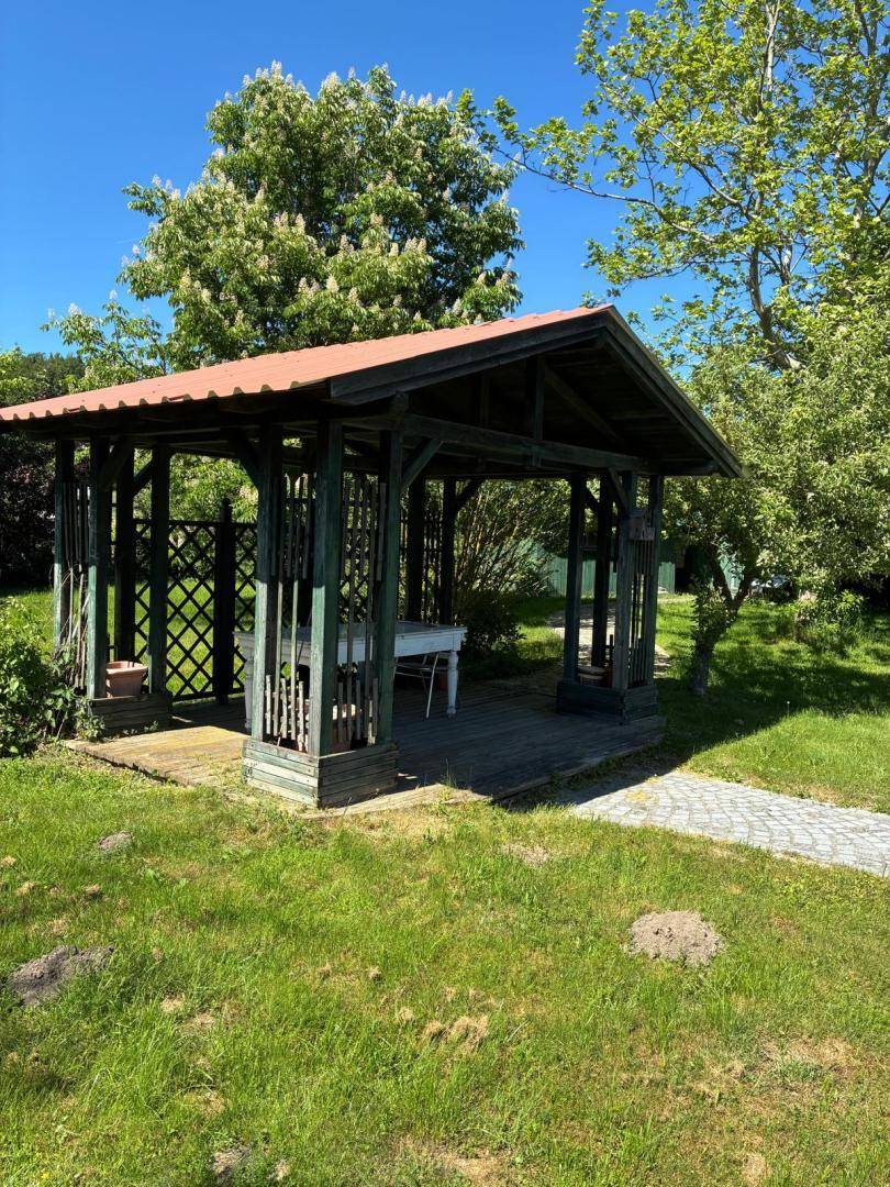 Attraktiver Holzpavillon mit Tisch, umgeben von grünem Rasen und Bäumen, zugänglich über einen gepflasterten Weg.