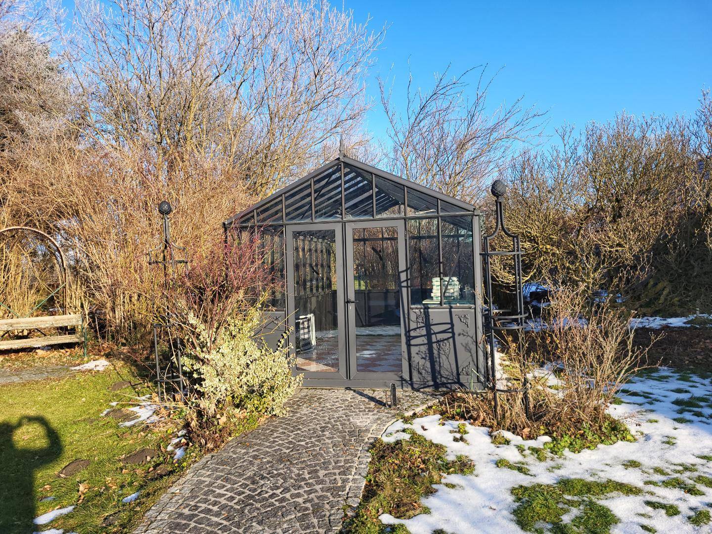 Modernes Gewächshaus mit gepflastertem Zugang, umgeben von kahlen Sträuchern und Schneeresten im Garten.