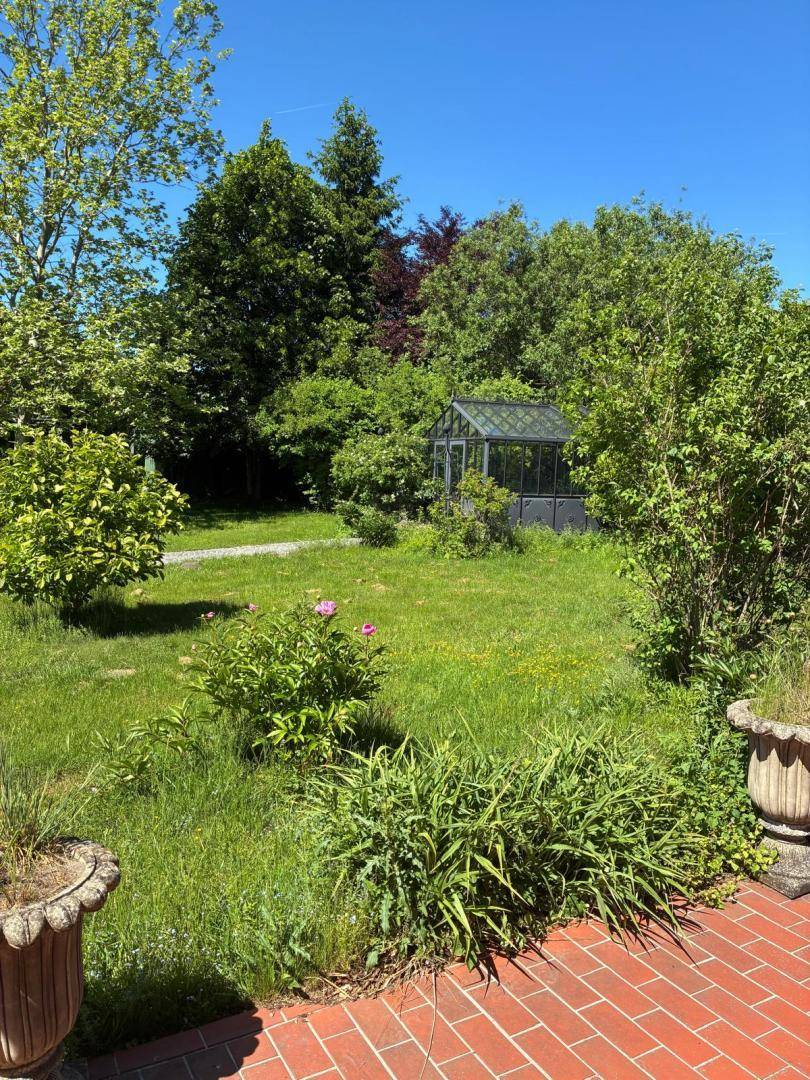 Blick vom Terrassenbereich in den grünen Garten mit üppigen Büschen und einem Gewächshaus im Hintergrund.