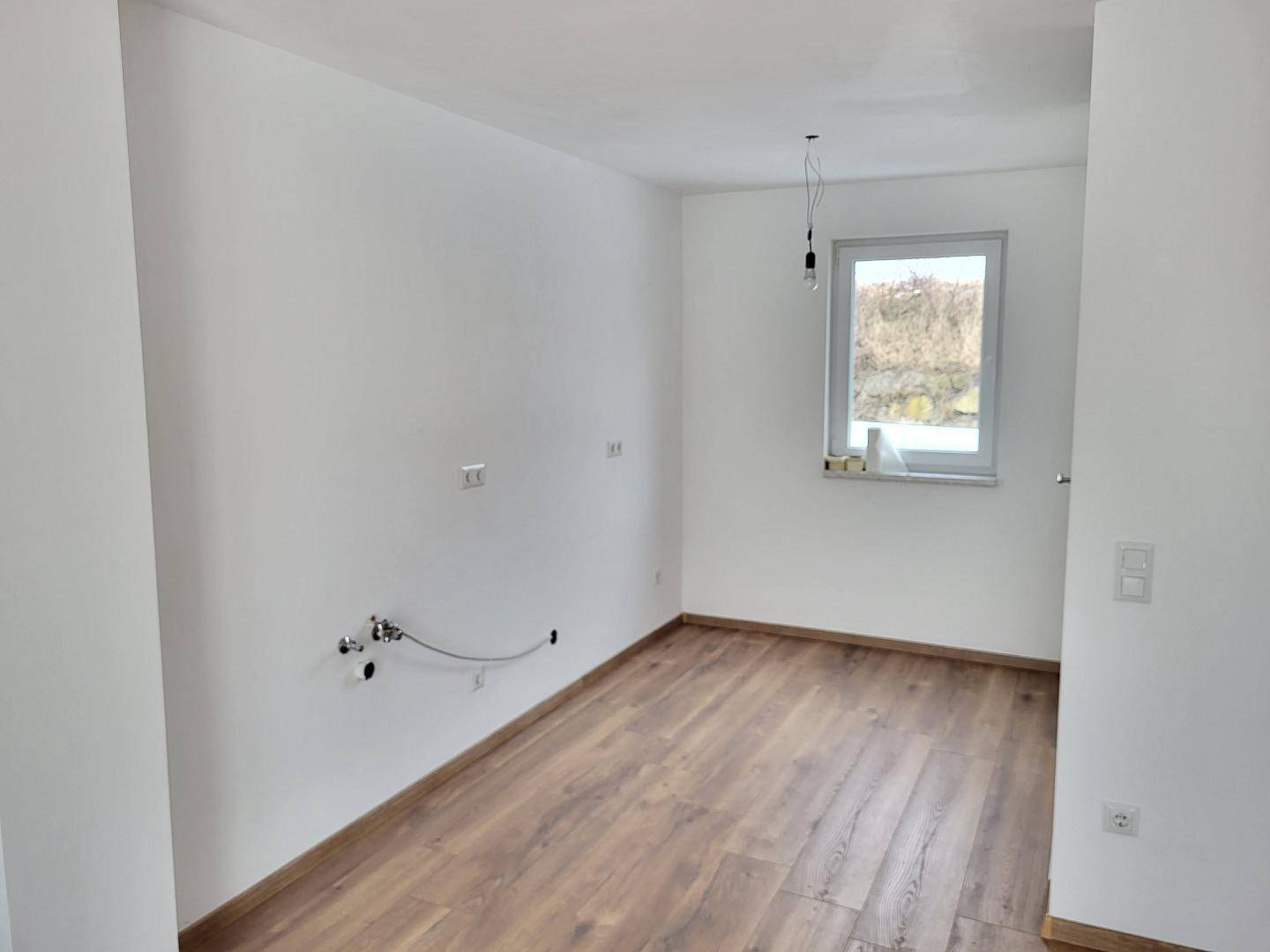 Leerer Küchenraum mit Anschlüssen für eine zukünftige Einbauküche und Fenster.
