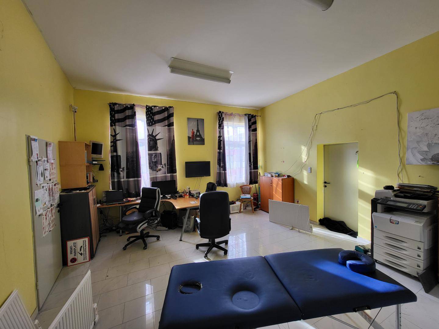 Geräumiges Büro mit mehreren Arbeitsplätzen und einer Massageliege, gelbe Wände.