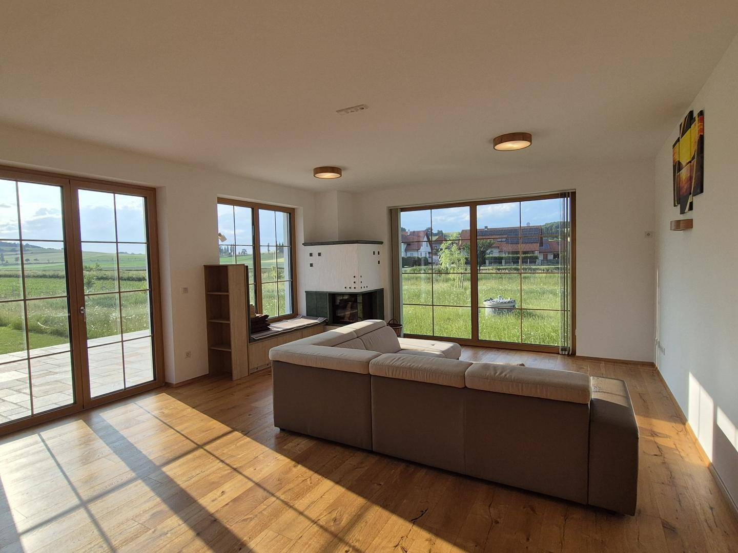Helles Wohnzimmer mit Holzboden, Kachelofen und großen Fenstern mit Landblick.