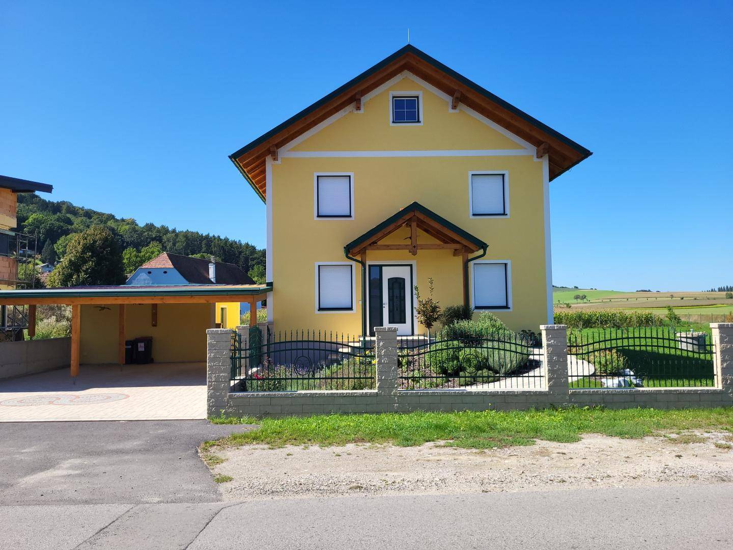 Vorderansicht des gelben Einfamilienhauses mit Carport, Zaun und Blick auf die Landschaft.