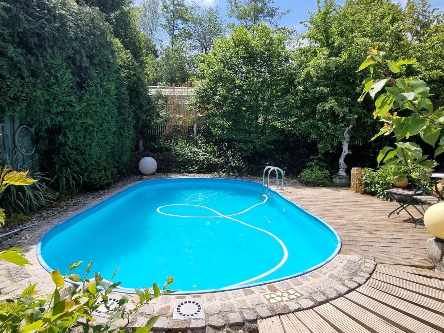 Blauer Swimmingpool mit Holzdeck und üppiger Bepflanzung im sonnigen Gartenbereich.